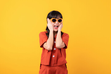 Portrait young beautiful asian woman happy smile dressed in orange clothes and sunglasses looking surprised, reacting amazed, raising eyebrows impressed isolated on yellow studio background.