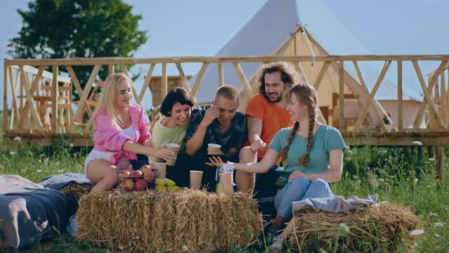 Campsite very charismatic and attractive ladies and guys have fun time together they drinking and looking over the smartphone watching something together while sitting down on the haystack