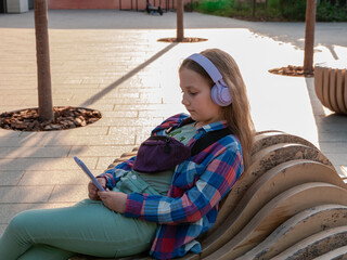 Blonde child purple headphones checkered shirt listen to music podcast using smartphone bench outside city street urban lifestyle. Concentrated young girl online audio book Mental health Stress relief