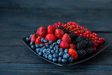  Ripe berries of strawberries, blueberries, blackberries and red currants on a square black plate on a wooden background
