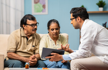 Indian doctor explaining from digital tablet to senior couple about medical report at home -...