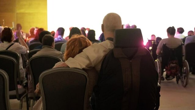 A disabled person is sitting in a wheelchair in a concert hall and watching a gig with his wife. Diversity