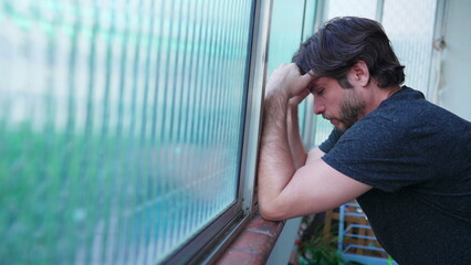 Stressed man struggling with mental illness leaning by window suffering from emotional despair. One...