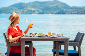 Woman in hat enjoys breakfast with sea view at luxury hotel resort on tropical beach. Girl savoring tasty food in beachfront cafe with personal watercraft on background. Vacation, sea holidays concept