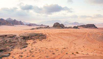 Fototapeta na wymiar Orange red sand desert, rocky formations and mountains background, overcast sky above, camp tents visible at distance - typical scenery in Wadi Rum, Jordan