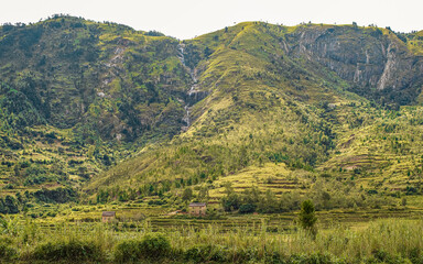 Typical Madagascar landscape in region near Ambohimanjaka. Terrain with small rocky hills covered with bushes and few trees, red clay houses, rice fields in foreground, waterfall at distance