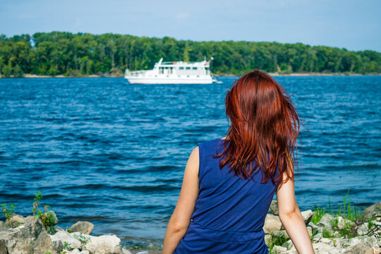 A traveler girl with red hair looks at a ship in the river from behind