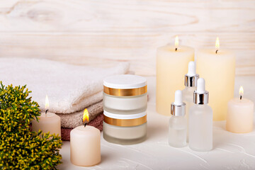 Obraz na płótnie Canvas Cosmetic jars with cream and white glass dropper bottles among burning candles. Towels and plant complete spa concept. Care and relaxation, beauty and health. Selective focus
