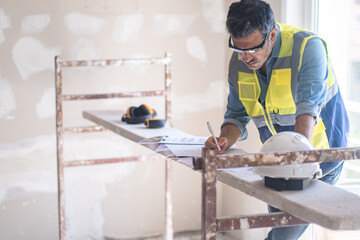 Focused architect writing notes on project documentation stands near construction safety equipment professional specialist in vest works with papers before renovation process