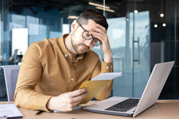 Shocked young man businessman sits in the office at the table and reads the received letter, documents. He holds his head worriedly - Powered by Adobe