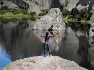 middle-aged woman with her back turned and arms outstretched in front of a rock in a pond in the middle of a rocky forest