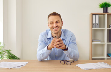 Portrait of happy businessman or remote worker at online business meeting via video conference call. Young man in shirt sitting at office table, holding pen, looking at camera and smiling