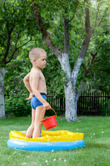Active little boy plays in rubber pool with red bucket on backyard.