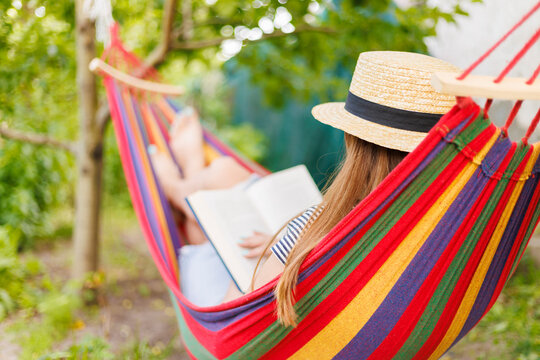 Young woman reading book while lying in comfortable hammock at green garden