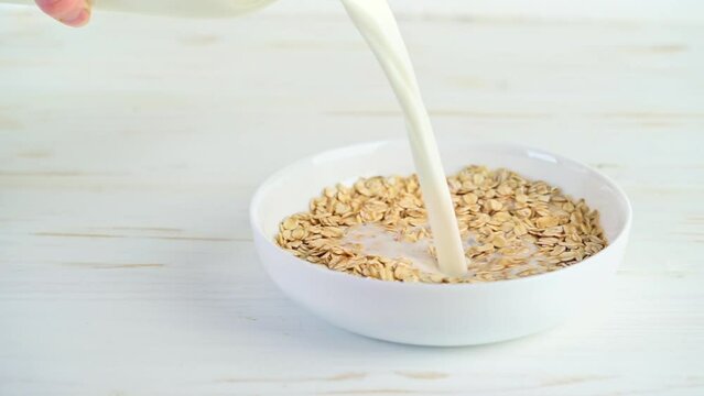 Milk pouring into oat flakes in a bowl on white wooden background. Slow motion