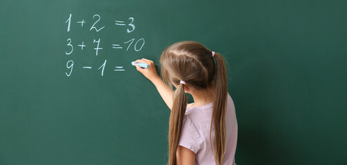 Cute little schoolgirl writing on blackboard during math lesson - obrazy, fototapety, plakaty