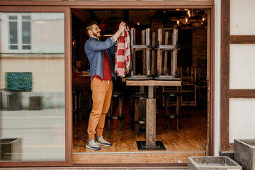 Fototapeta premium Happy Caucasian waiter opening the door at a cafe and preparing a venue in the morning.