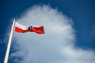 Flaga Powstania Warszawskiego, wyraźnie wywieszona na maszcie, symbolizująca wytrwałość i narodową dumę na tle jasnego nieba.