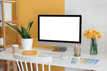 Modern PC, beautiful narcissus flowers, houseplant, coffee, notebook and paint color palettes on table near color wall