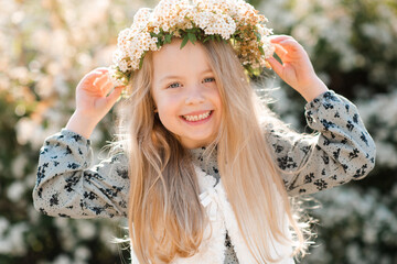 Smiling child girl wear floral wreath and dress posing over blooming tree outdoor at nature background. Looking at camera. Childhood. Springtime.