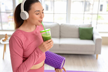 Sporty young woman in headphones drinking vegetable juice at home