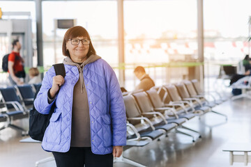 Middle aged woman in eyeglasses with hand luggage, backpack waiting by the boarding gate at plane of the airport with a smartphone in her hands. Active adventure retirees lifestyle travel.