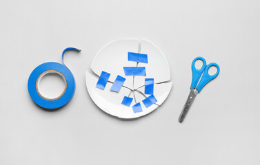 Broken plate with adhesive tape and scissors on light background