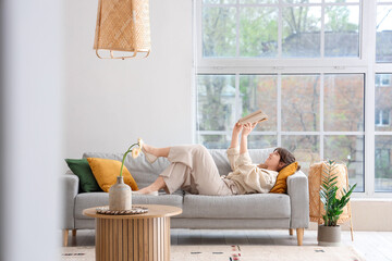 Happy young woman lying on grey sofa and reading book in interior of light living room, view from hallway