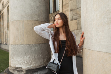 Fashionable beautiful young caucasian girl model with bag in a stylish black dress with a white shirt walks near a vintage building with columns