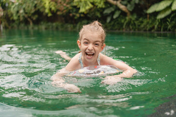 Happy little girl playing with inflatable ring in outdoor swimming pool on  summer day. Preschool child learn to swim. Kid play in tropical resort with toys. 