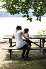 Rear view of mother and daughter sitting on a picnic table outside by a river. 