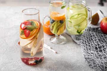 Glass, mason jar and bottle of infused water with different sliced fruits on grey table