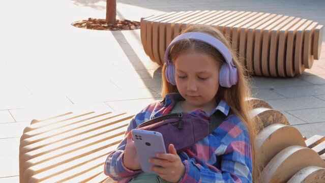 Blonde child headphones checkered shirt listen to music podcast smartphone outside city street urban lifestyle. Concentrated girl online audiobook social media. Mental health application Stress relief