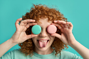 Headshot portrait of funny cute young ginger girl with curly hair holding tasty macaroon sweet...