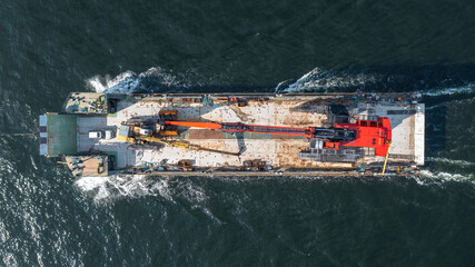 Barge loaded with tracked harbour material handling cranes, being pulled across the open ocean. Aerial top down view.