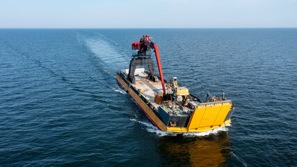 Obraz premium Yellow barge loaded with tracked harbour material handling cranes, being pulled across the open ocean. Aerial front view.