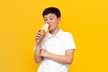 asian little boy of ten years old licks ice cream cone on yellow isolated background