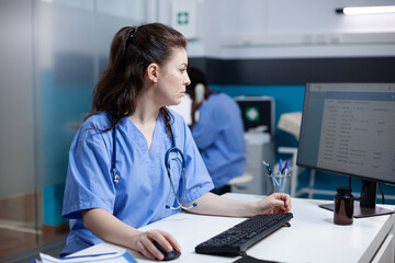 Caucasian nurse checking appointments list on computer monitor, sitting at clinic desk. Focused adult woman healthcare expert working in medical office with stethoscope around neck - obrazy, fototapety, plakaty
