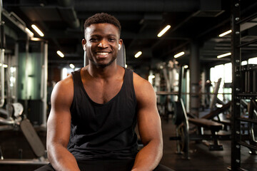 young athletic african american man sits in dark gym and smiles, athletic guy rests in fitness club
