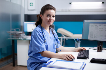 Smiling caucasian nurse inside modern, clean, clinical office, sitting at desk. Young cheerful professional adult woman employee wearing scrubs and stethoscope, looking at camera, portrait