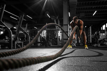 athletic african american man trains with ropes in dark gym, motivated guy waving ropes in fitness...