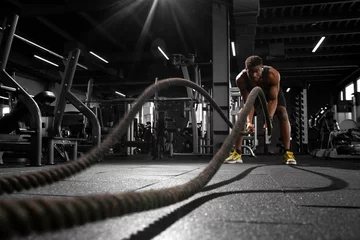 Fotobehang Sportschool athletic african american man trains with ropes in dark gym, motivated guy waving ropes in fitness club  © Богдан Маліцький