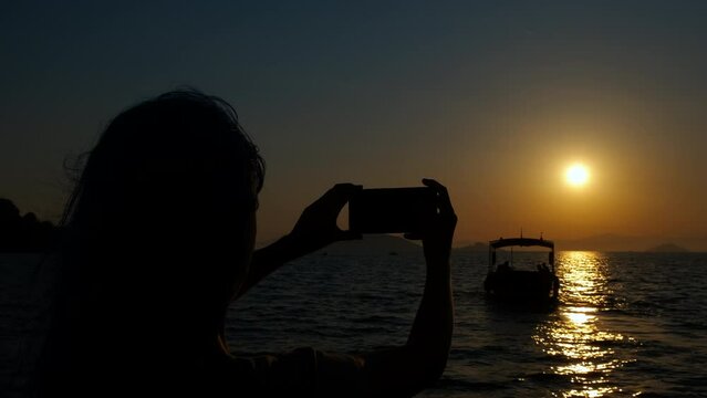 Teen silhouette use smartphone by sea sunset. Silhouette of girl with phone taking photos of orange sun during sunset on the bay.