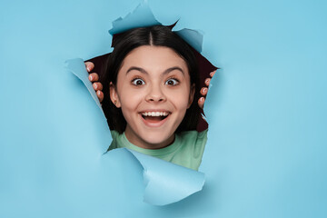 Excited young latin teenage girl looking at camera over torn blue background. Beautiful young female