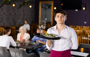 Foto op Canvas Young waiter guy is standing with order tray in spacious guest room in restaurant. Male restaurateur smiles and looks at camera. Cafe patrons eating in background. © JackF