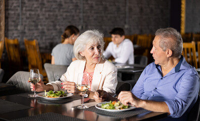 Fototapeta na wymiar Cheerful elderly couple spending time together in cozy restaurant. Man and woman having fun while talking and enjoying light dinner with wine at table