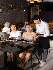 Young male waiter in uniform takes order from two women in restaurant