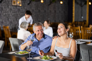 Smiling senior man spending time with young girl in restaurant. Carefree couple laughing merrily, enjoying delicious food and drinking wine at dinner. Age-gap relationships concept