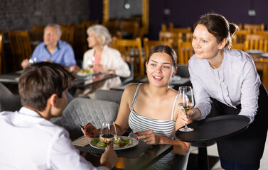 Polite smiling waitress serving ordered drinks to young couple sitting at table, visiting restaurant for romantic dinner