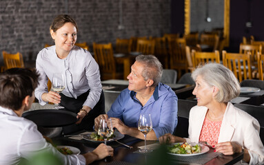 Elderly man and woman sitting in cafe and chatting sweetly with adult son. Waitress girl serves glass of wine to guest, visitors eat salad